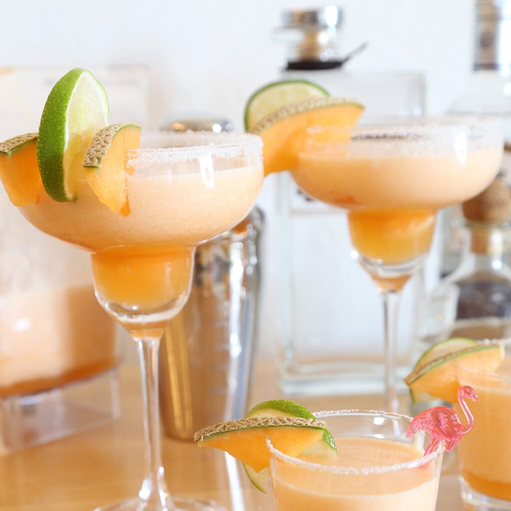 Two cantaloupe margaritas in margarita glasses and two are in cocktail glasses decorated with a couple slices of cantaloupes and lime. A shaker and bottles behind the glasses.