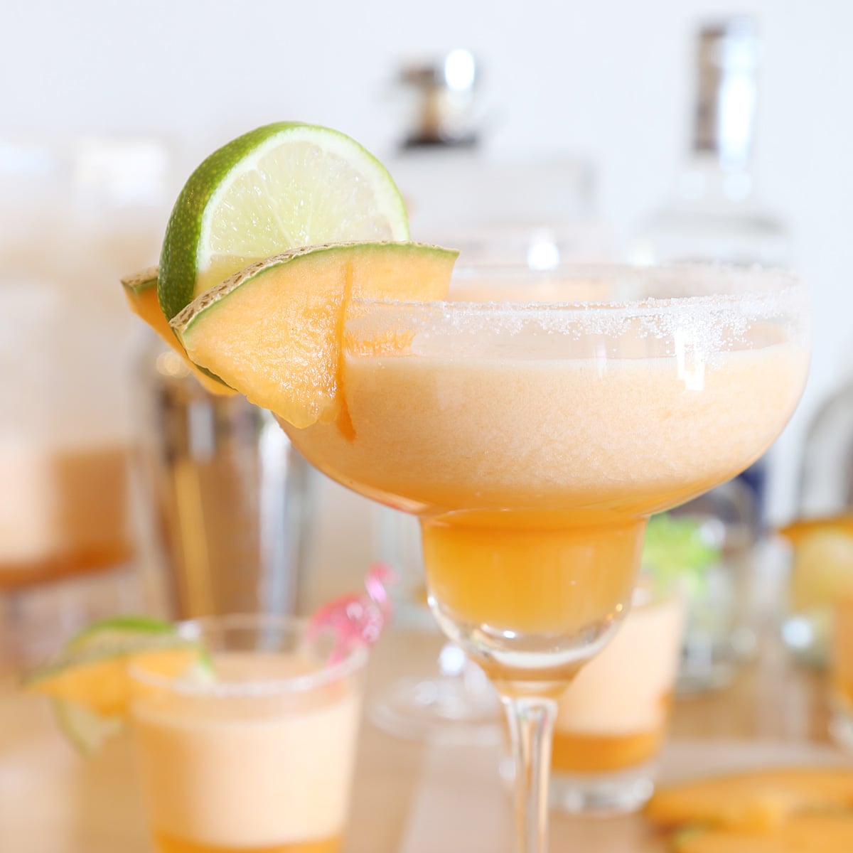 One cantaloupe margarita in a margarita glass and two are in cocktail glasses decorated with a couple slices of cantaloupes and lime. A shaker and bottles behind the glasses.