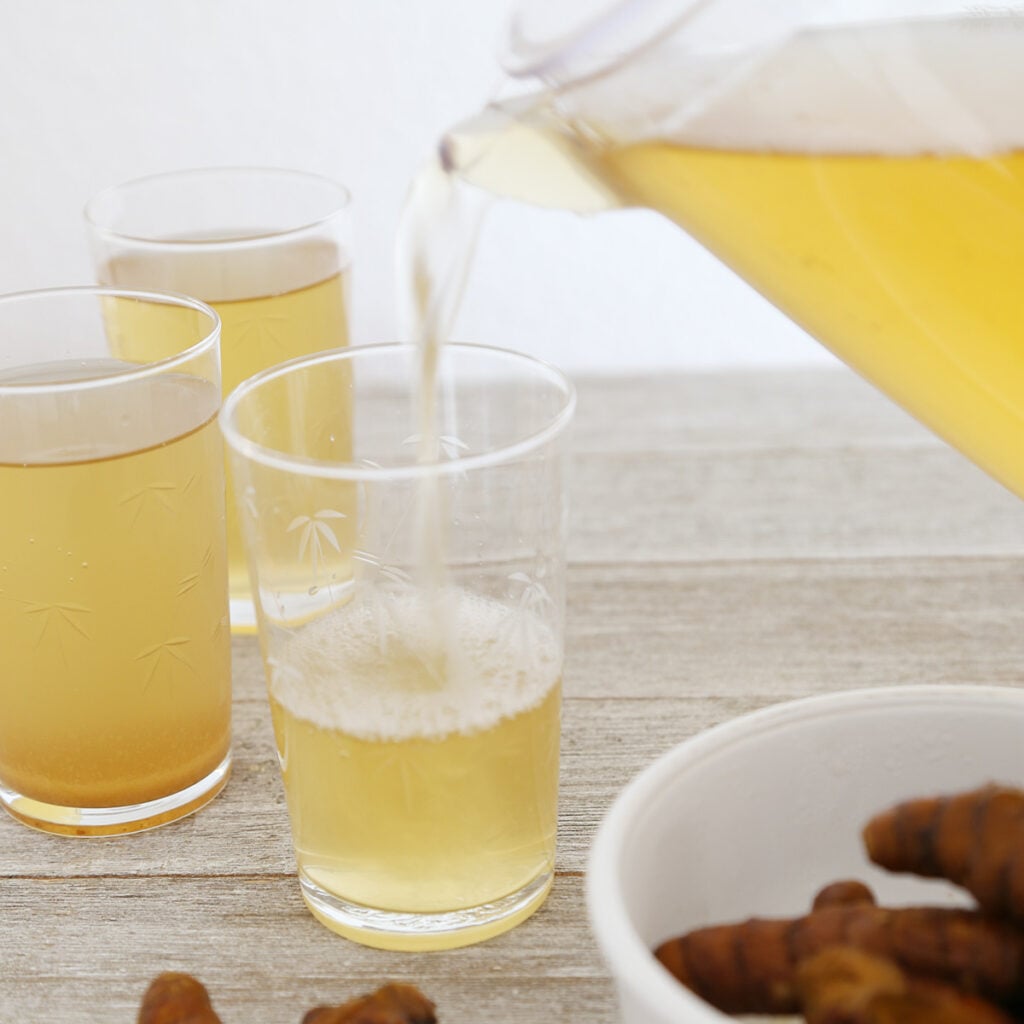 A pitcher of cold turmeric tea poured into 3 glasses and some whole unpeeled turmerics in a bowl.