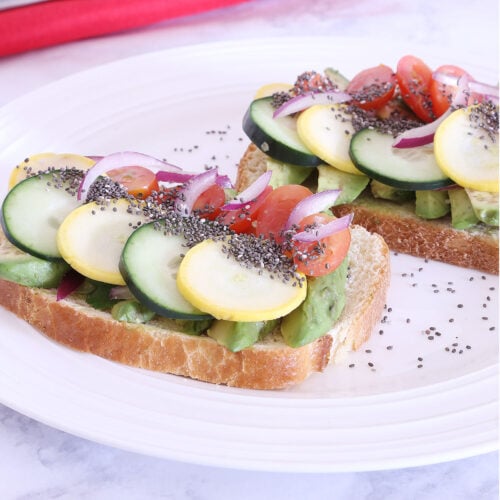 Two slices of toasted bread topped with cucumber, tomato, avocado, red onion, zucchini sprinkled with chia seeds on a plate.