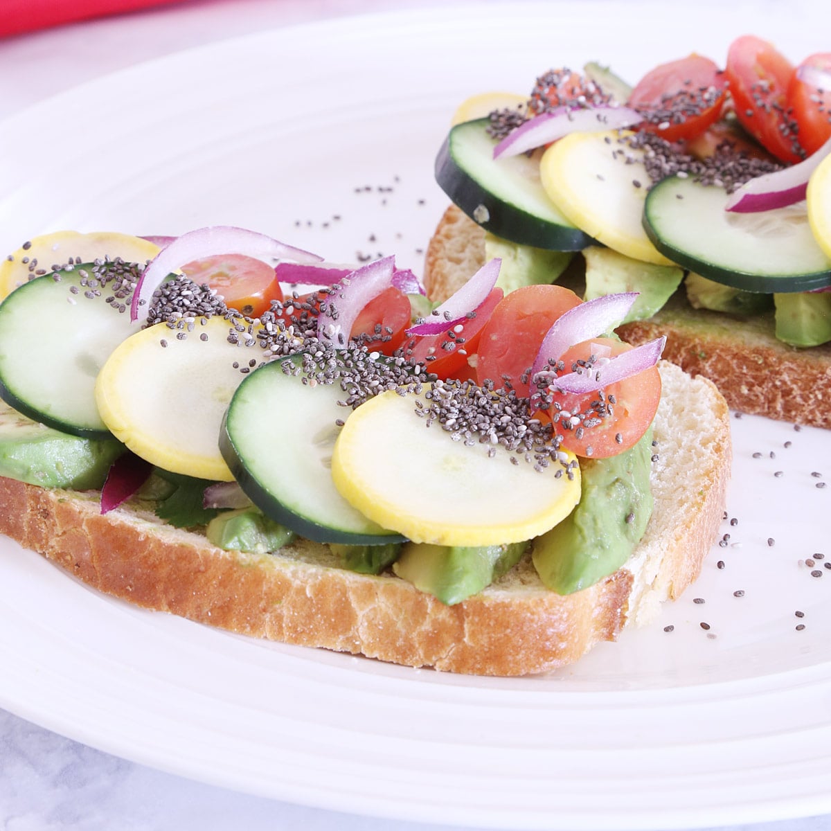 Two slices of toasted bread topped with cucumber, tomato, avocado, red onion, zucchini sprinkled with chia seeds on a plate.