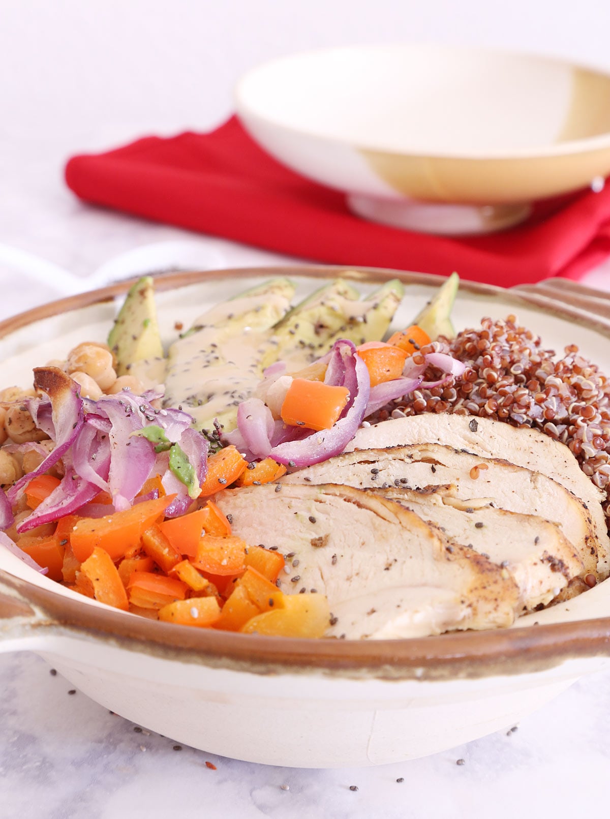 Chicken and Quinoa Bowl has roasted onion, red bell pepper, garbanzos, sliced avocado, lemon pepper chicken, chia seeds drizzled with creamy tahini dressing. The bowl cover sits on a red napkin in the background.