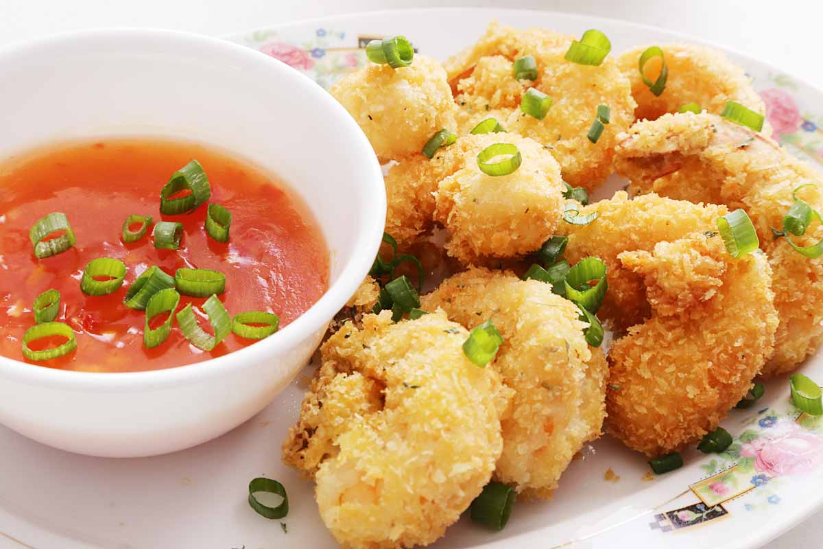 Eight crispy breaded shrimp on a platter with a side of sweet and sour sauce on a small bowl, and both are garnished with green onions.