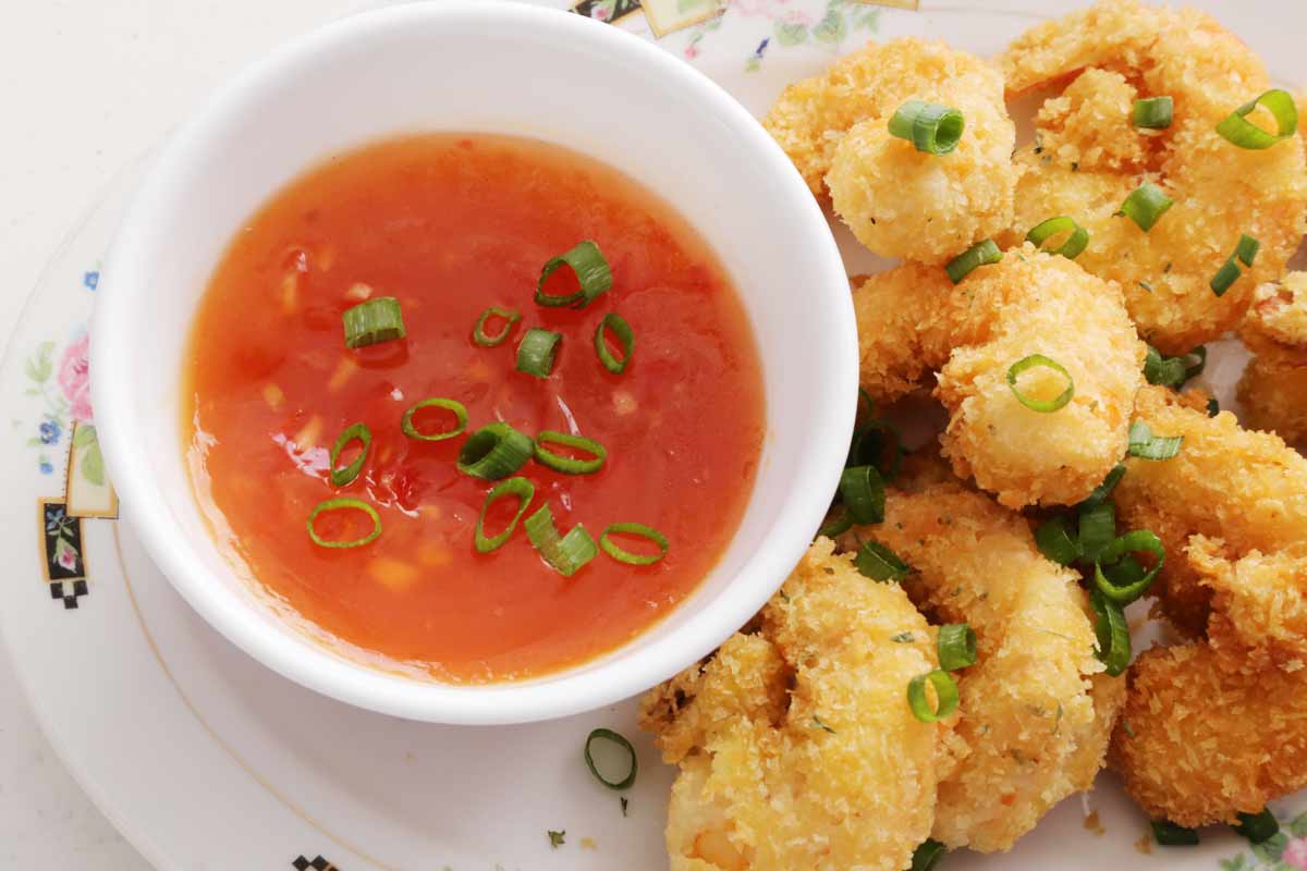 Eight crispy breaded shrimp on a platter with a side of sweet and sour sauce on a small bowl, and both are garnished with green onions.
