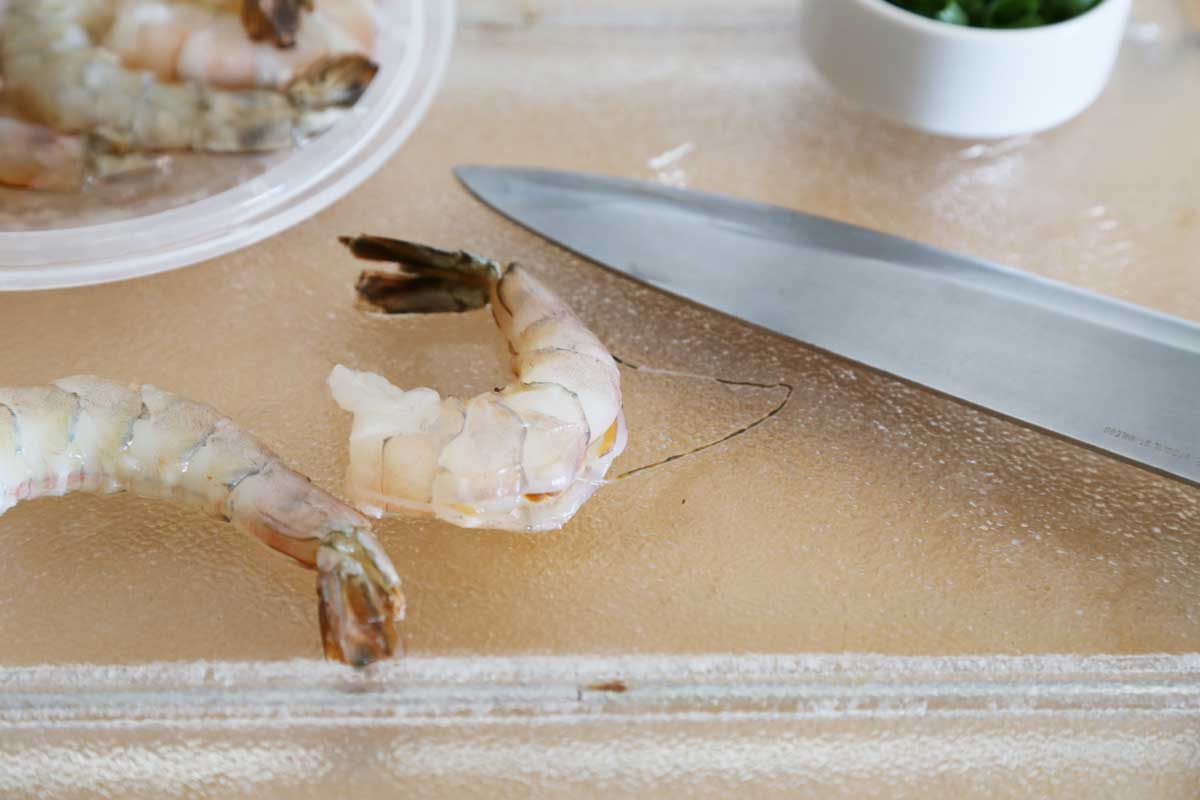 A shrimp is deveined with a knife on a cutting board. More shrimp on a plate and chopped green onions on a small bowl.