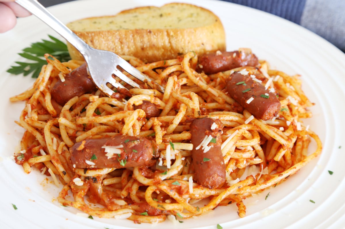 Spaghetti with whole little smokies sausages garnished with grated cheese and chopped parsley and a side of toasted garlic bread. A fork to pickup spaghetti.