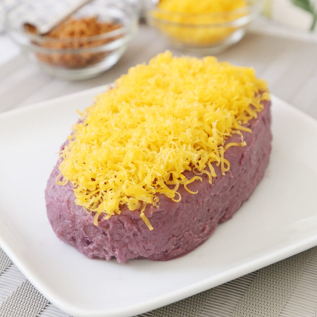 Ube halaya, a Filipino purple yam (ube) dessert, topped with grated cheddar cheese on a plate and a side of toasted coconut curds (latik) in a small bowl with a small spoon and more grated cheese in a small bowl.